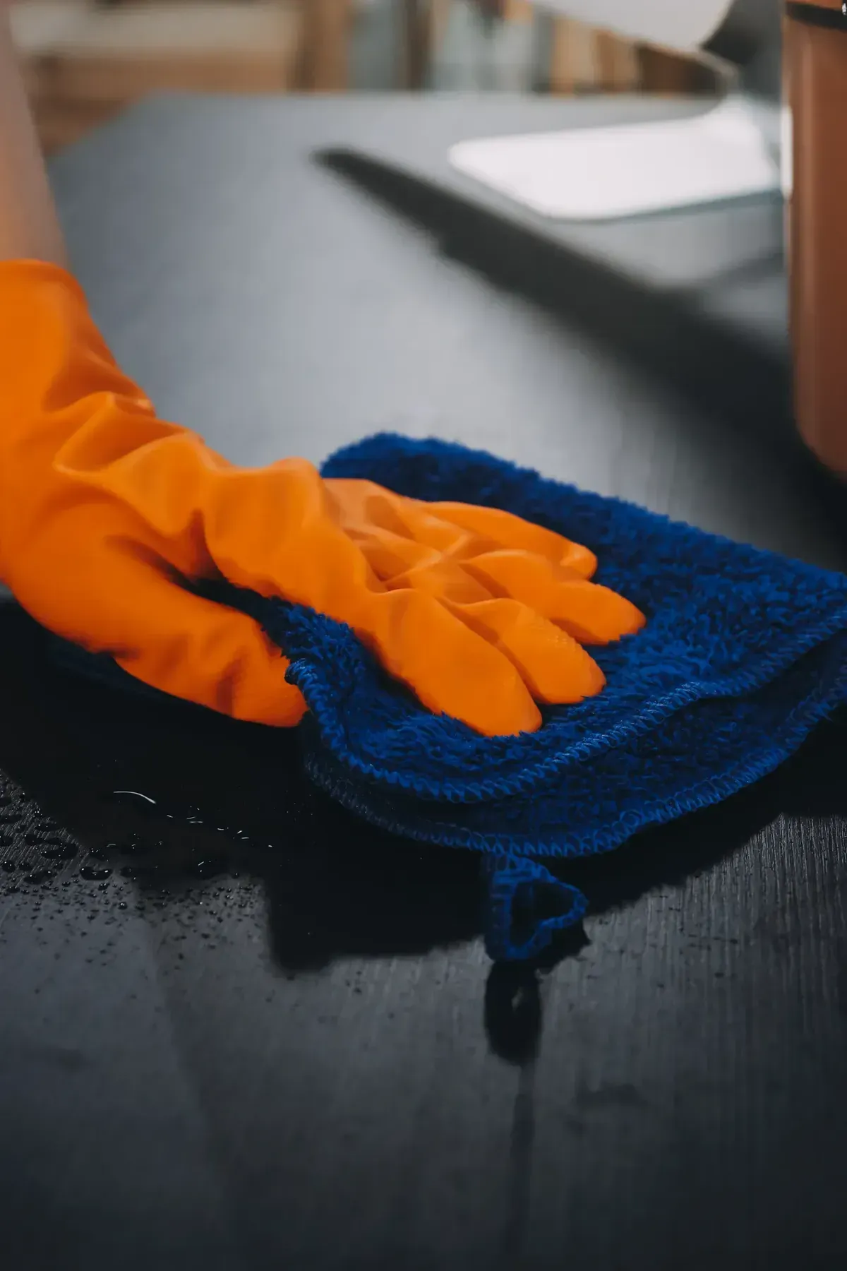 Mano enguantada de color naranja limpiando una encimera negra y húmeda con un paño azul.