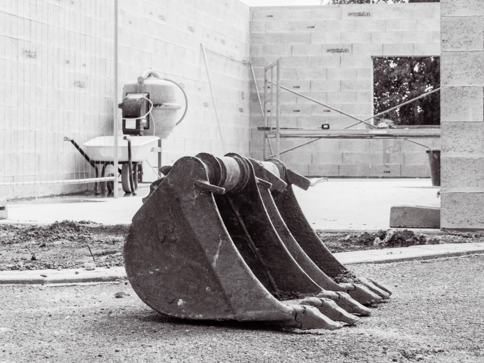 Godet d'excavatrice au premier plan, chantier de construction avec bétonnière et murs en arrière-plan.