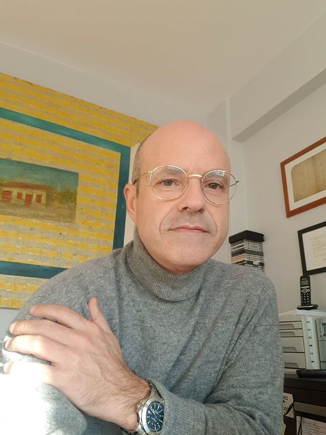 Un hombre calvo que lleva gafas y un suéter de cuello alto está sentado en una habitación con los brazos cruzados.