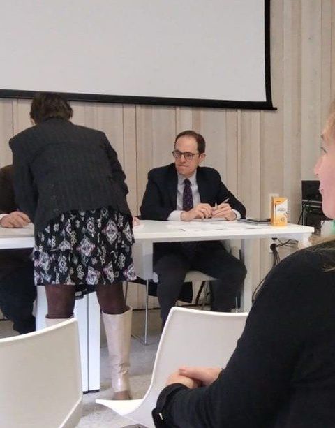 Un hombre con traje y corbata se sienta en una mesa frente a una pantalla de proyector.