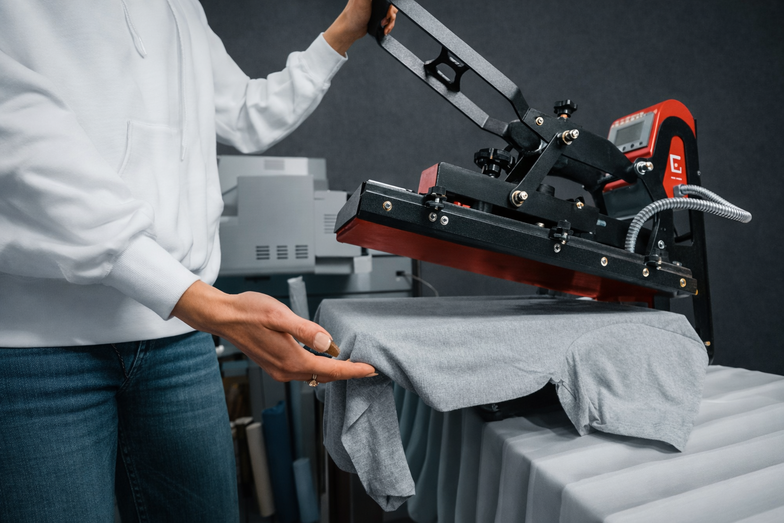 Una persona con una sudadera blanca se ajusta una camiseta gris debajo de una máquina de prensado térmico.