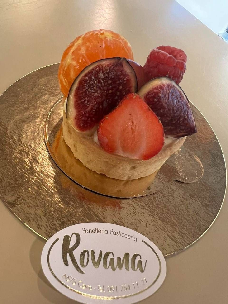 Crostata di frutta con fichi, lamponi, mandarini e fragole su un centrino dorato, con il logo 