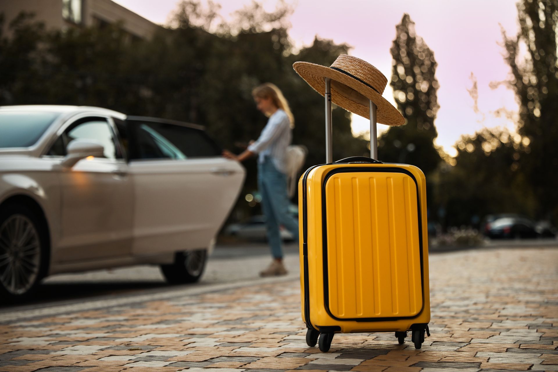 Une valise jaune avec un chapeau de paille et une femme ouvrant une portière de voiture.