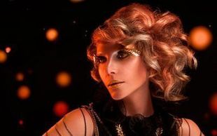 Una mujer con cabello rojo y maquillaje dorado está parada frente a un fondo negro.