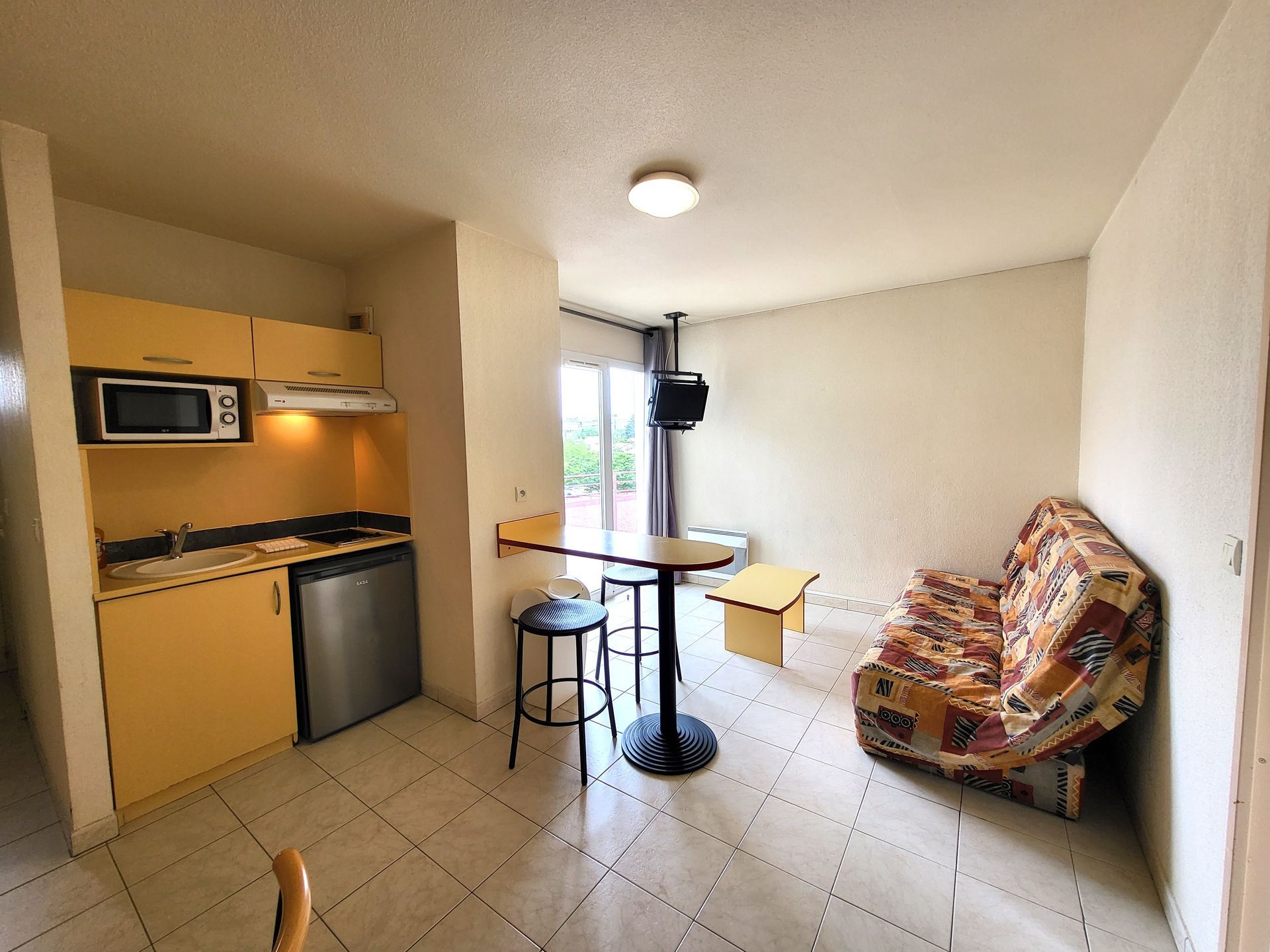 Petit studio avec kitchenette, table ronde et canapé à motifs près d'une fenêtre lumineuse