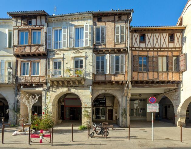 Immeuble historique à colombages avec arcades et balcons, situé dans une rue ensoleillée.