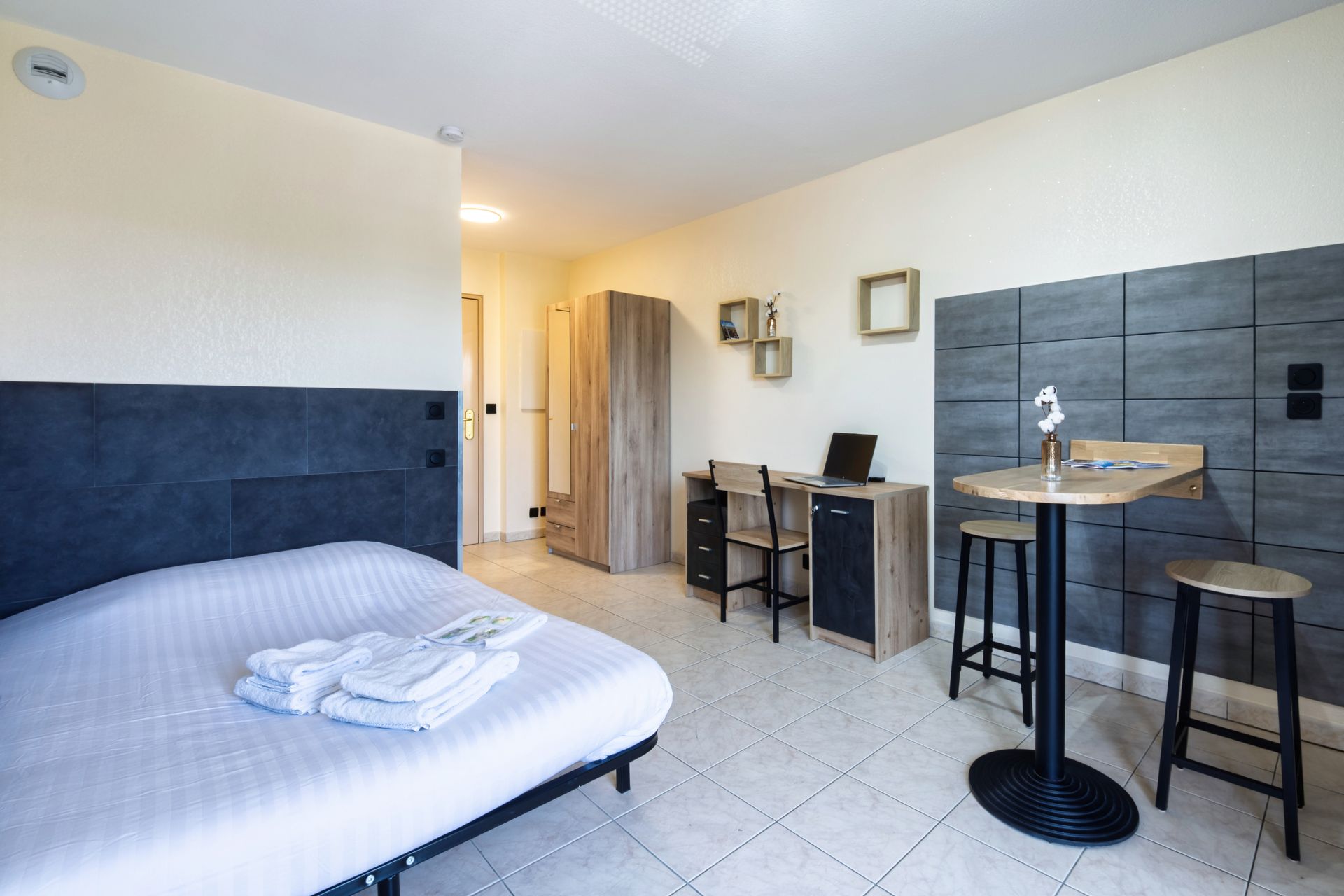 Petit studio avec lit, bureau, tabourets et kitchenette contre des murs carrelés