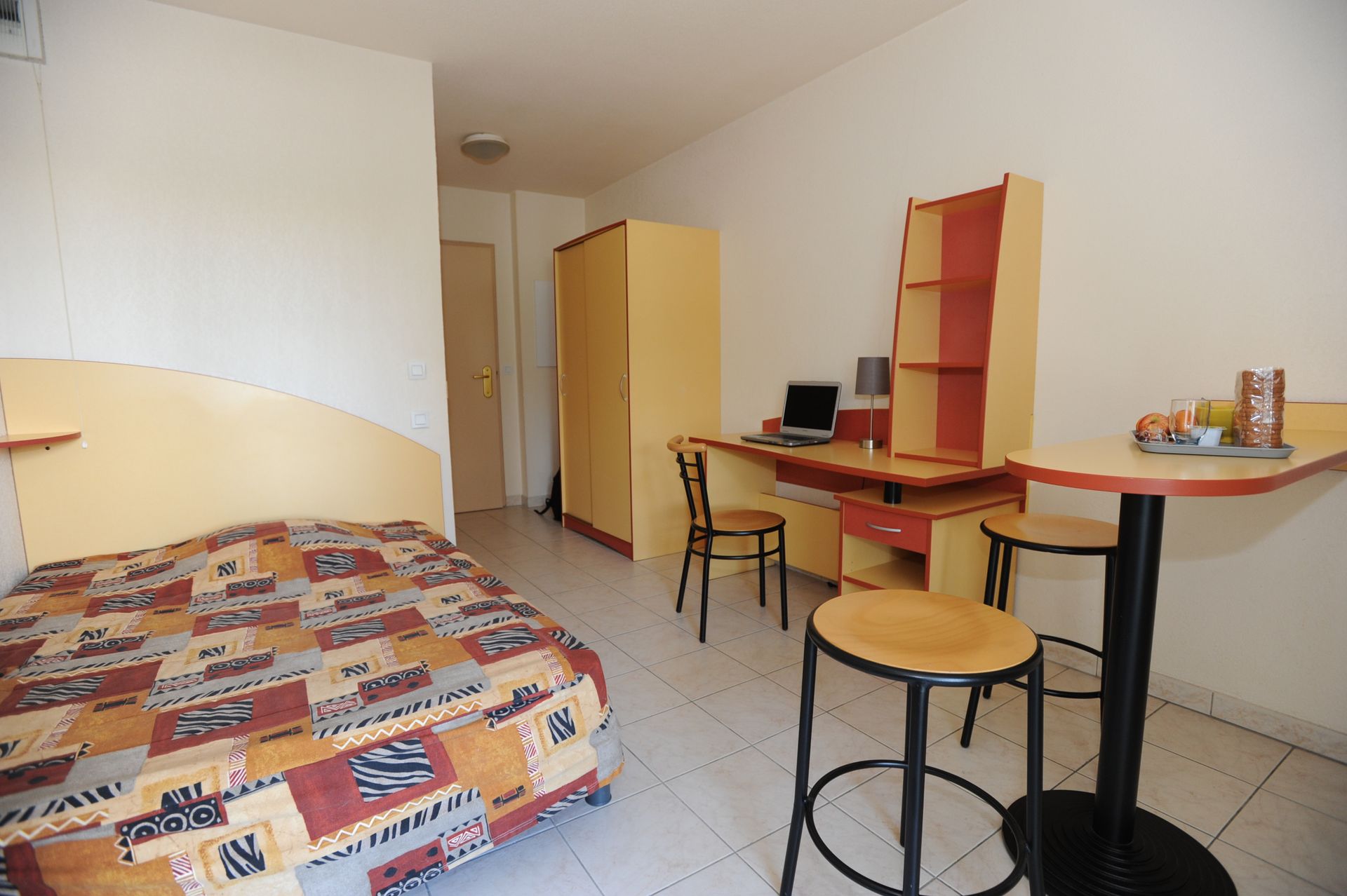Petite chambre meublée avec un lit simple, un bureau, une chaise, des étagères et des tabourets sur un sol carrelé.