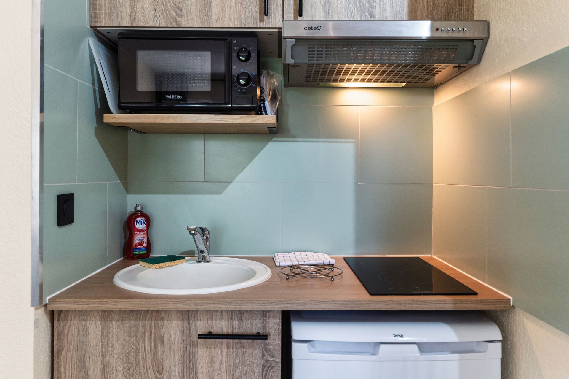 Petite kitchenette avec évier, micro-ondes, plaque de cuisson et mini-réfrigérateur sous des placards en bois clair.