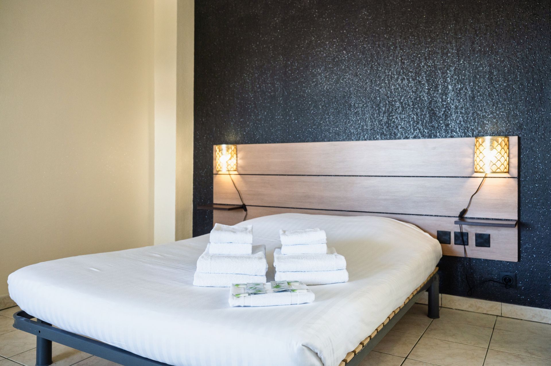 Lit d'hôtel impeccablement fait avec serviettes pliées dans une chambre moderne, éclairée par des appliques murales