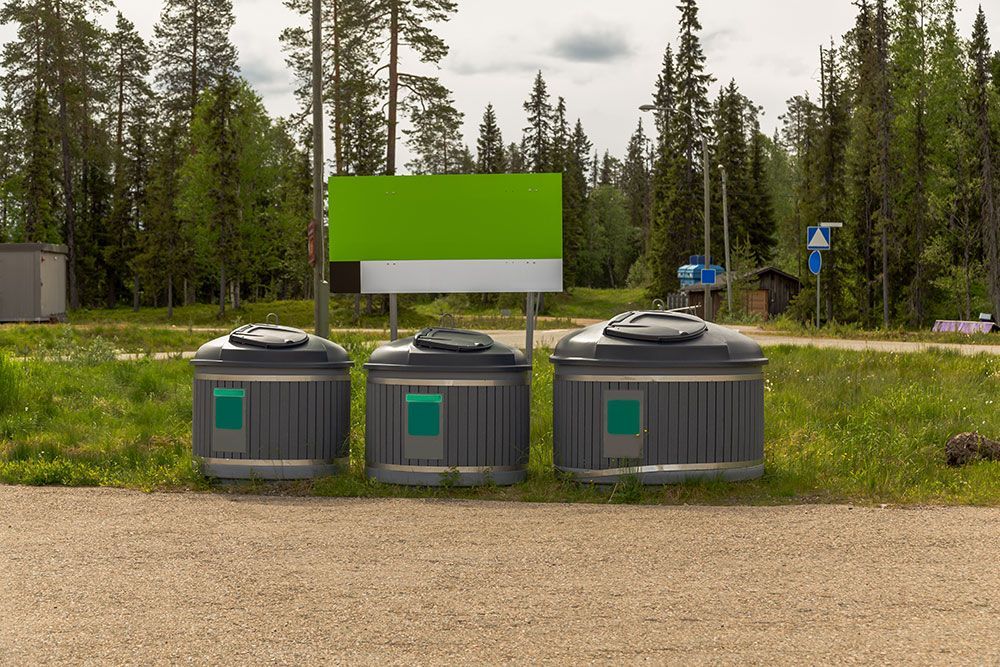 Kolme harmaata kierrätysastiaa kirkkaanvihreän kyltin vieressä metsäisellä ulkoalueella