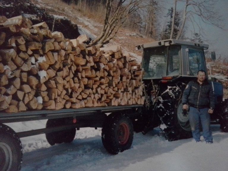 Un tracteur transportant une importante cargaison de bois de chauffage sur une route enneigée, avec une personne debout à côté.
