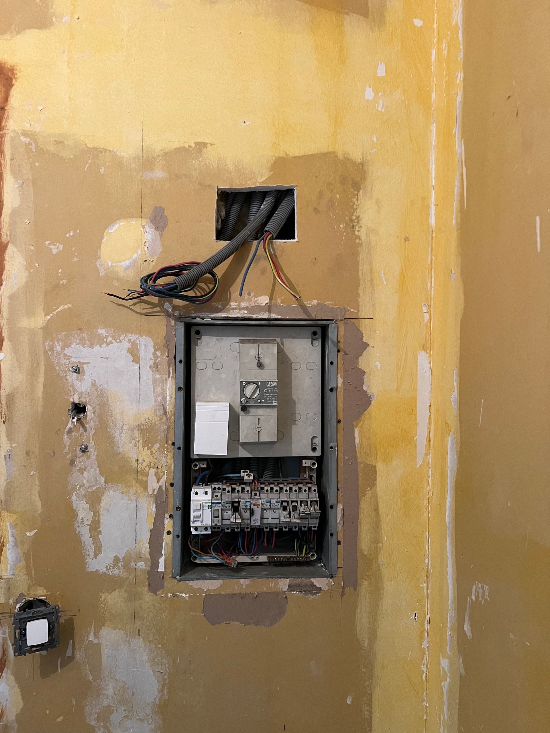 Tableau électrique ouvert dans un mur jaune délavé, avec fils apparents et plâtre écaillé.