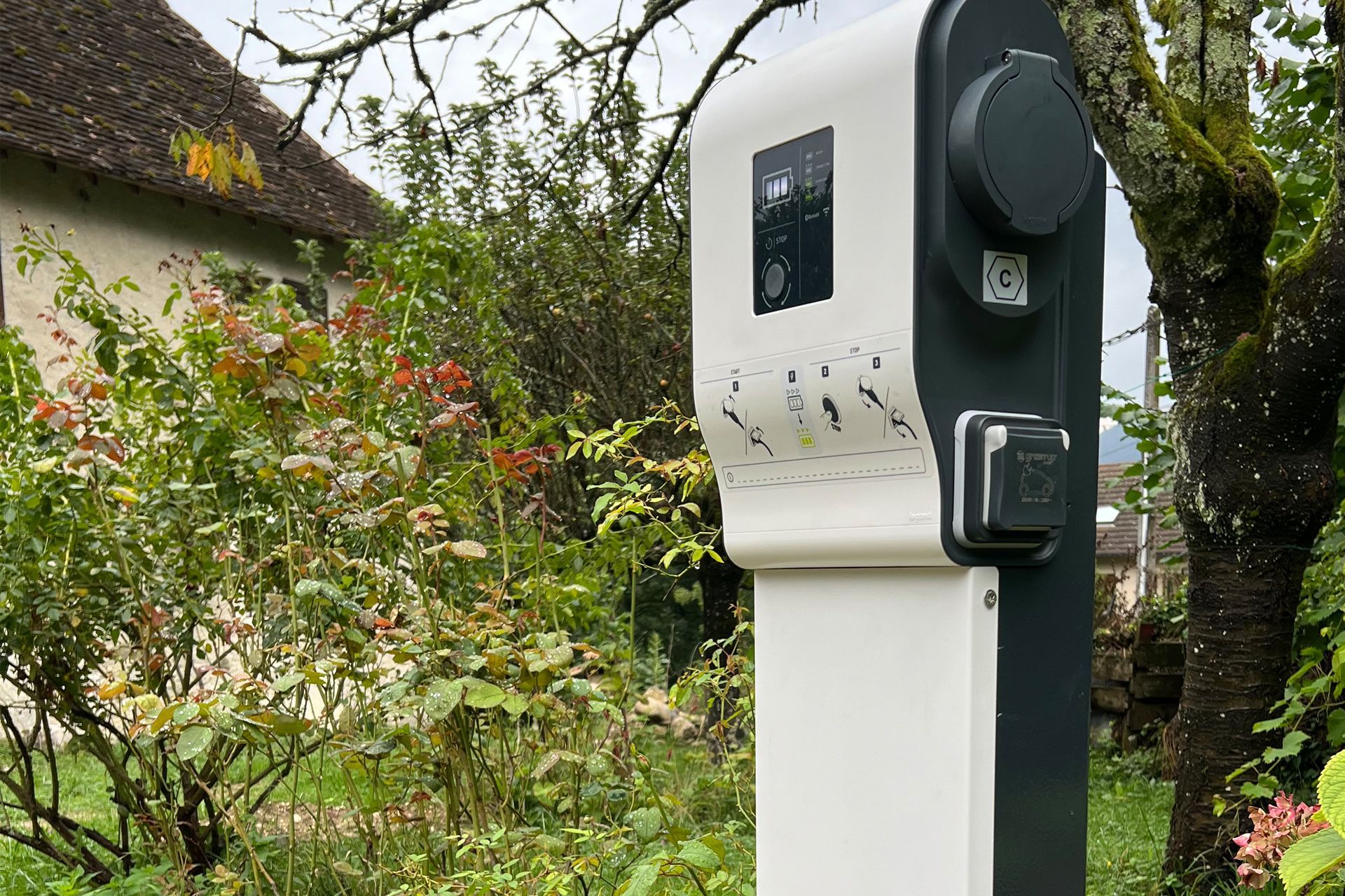 Borne de recharge blanche pour véhicules électriques située à côté d'un chemin de jardin bordé d'arbres et d'arbustes.