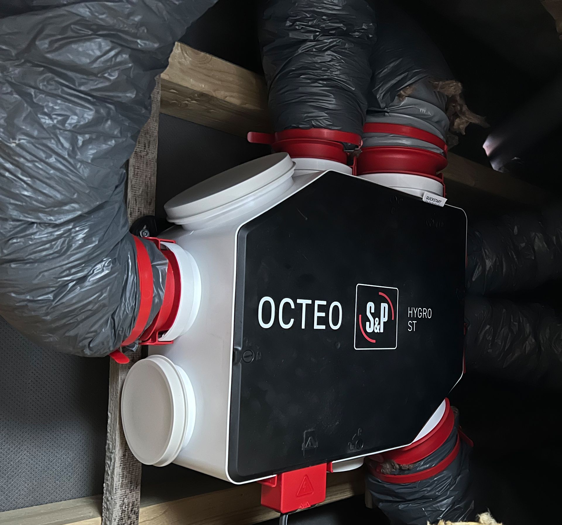 Dispositif OCTEO noir avec raccords de tuyau rouges, monté parmi des tuyaux isolés dans un espace utilitaire exigu.