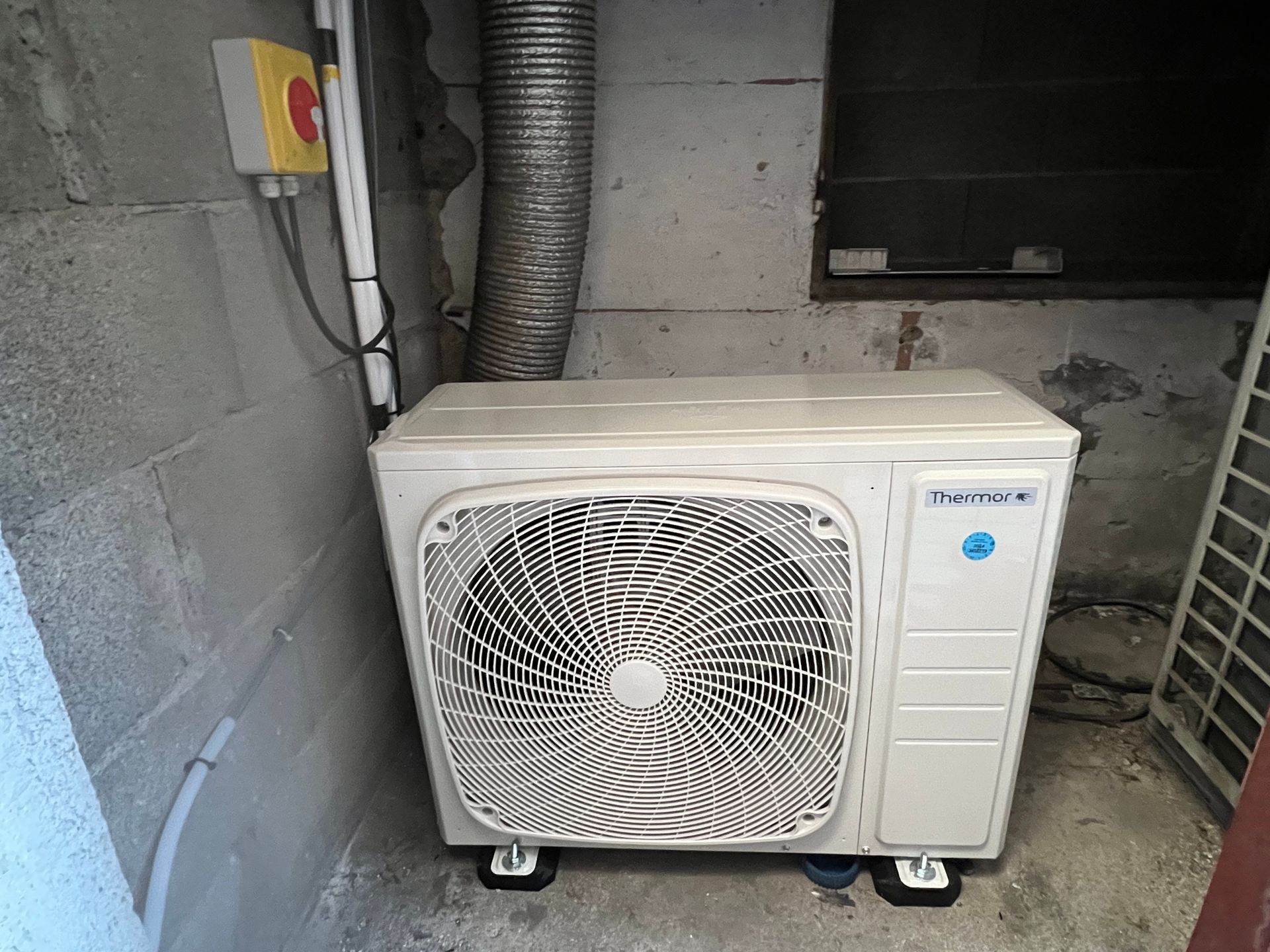 Unité de climatisation extérieure blanche sur un sol en béton dans un local technique en parpaings, avec une grille de ventilation et un tuyau d'évacuation.