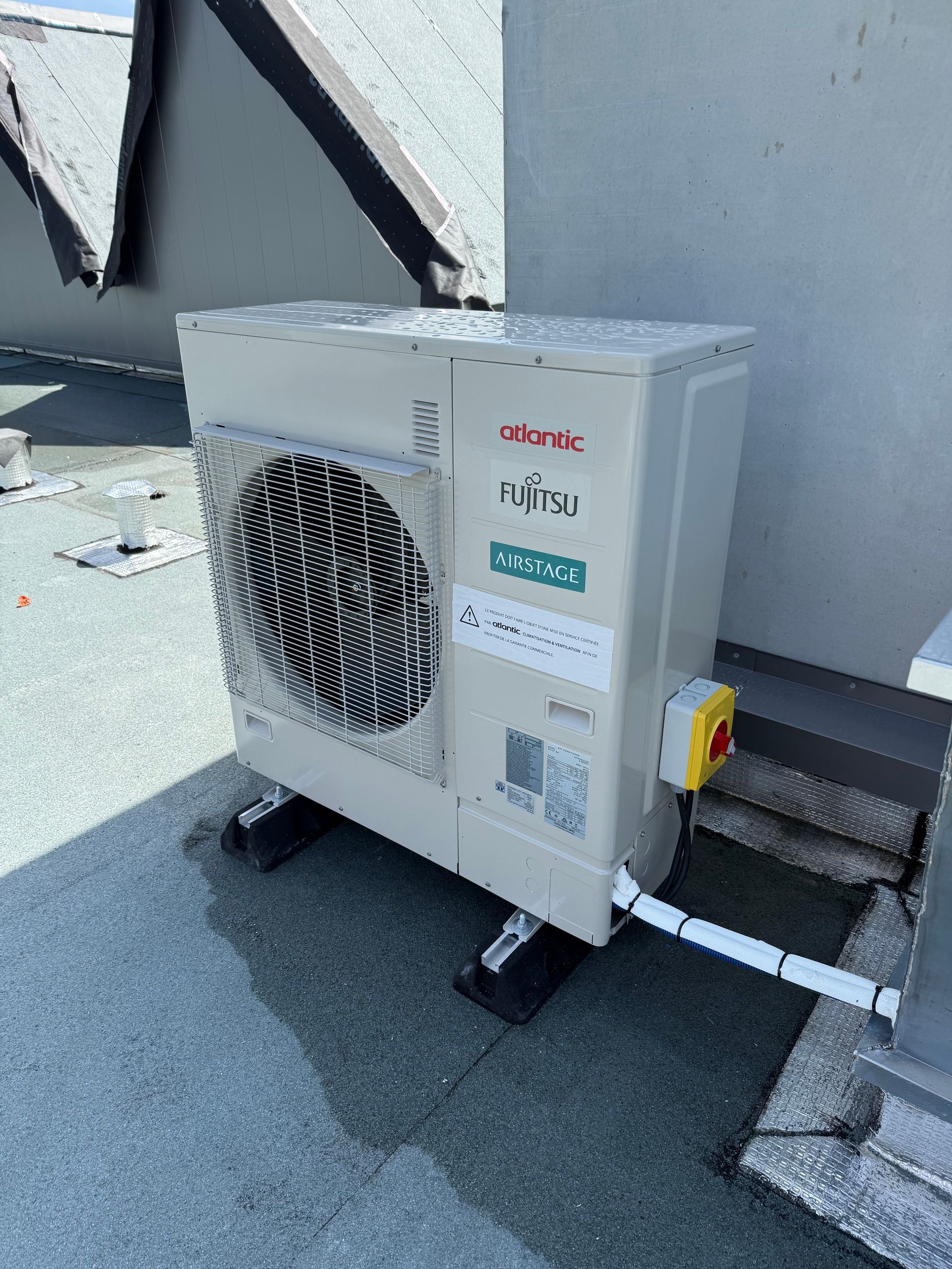 Unité de climatisation extérieure fixée à un mur sur un toit, avec des tuyaux et un boîtier de commande jaune.
