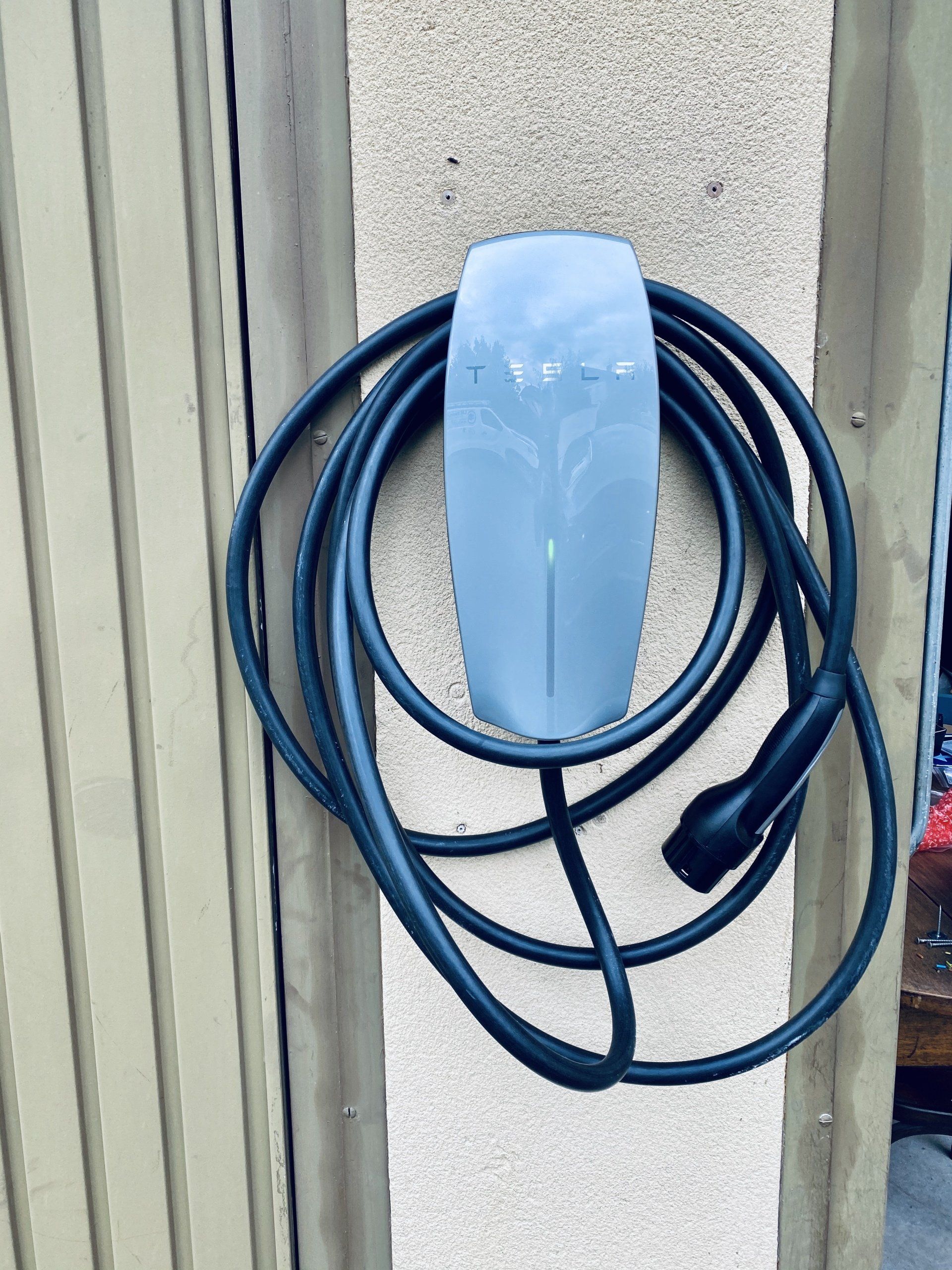 Borne de recharge électrique avec câble noir enroulé, suspendu à un crochet mural contre un mur extérieur beige.