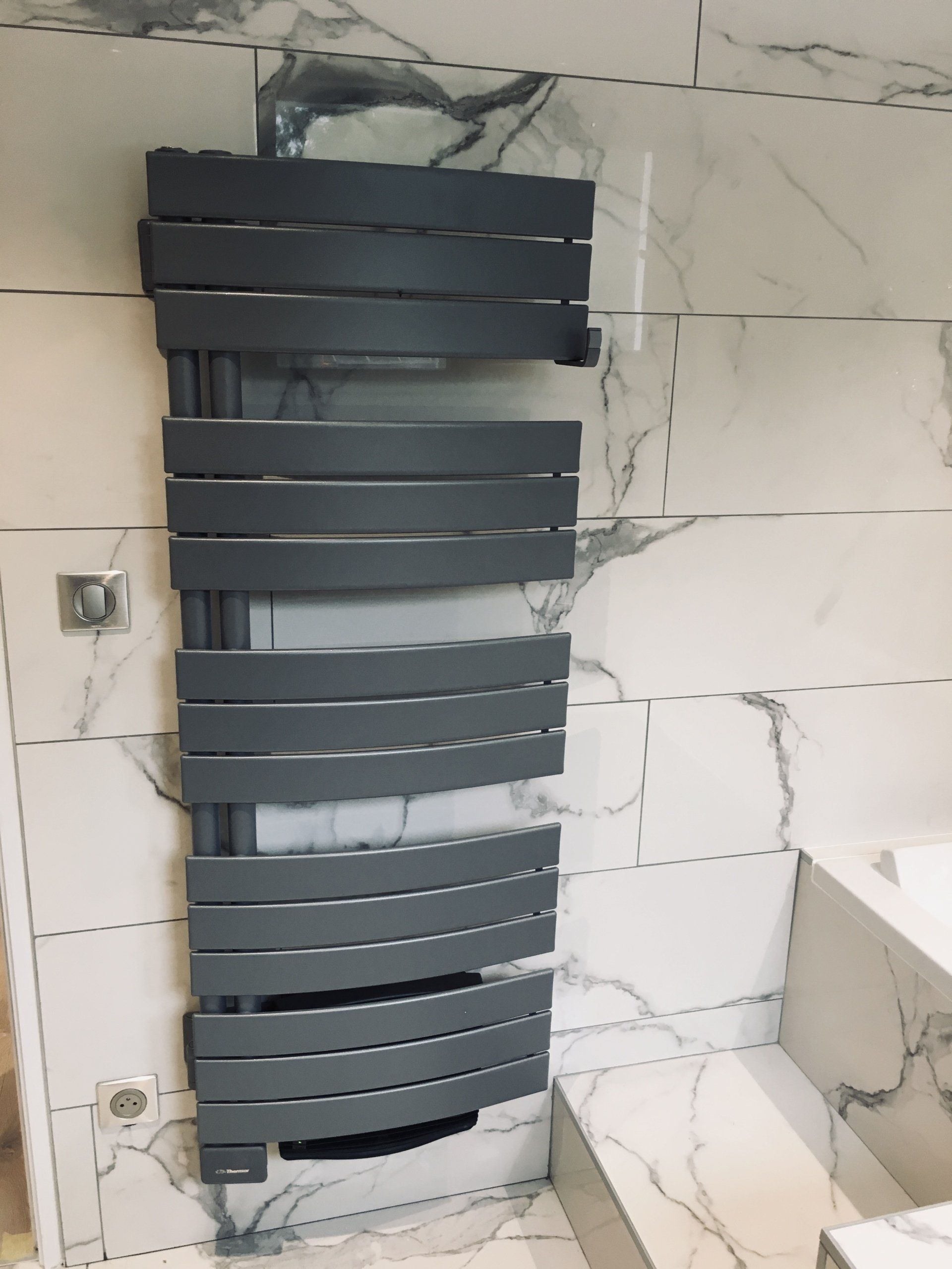 Radiateur sèche-serviettes noir sur un carrelage blanc à motif marbre dans une salle de bains, avec des moulures non finies à proximité