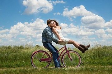 Junges Pärchen küssend auf einem Fahrrad in einem Feld vor einem blauen Himmel