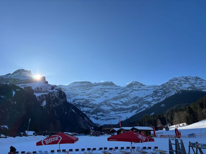Station de ski enneigée au coucher du soleil, avec des sommets montagneux, un ciel bleu et des parapluies rouges sur la neige.