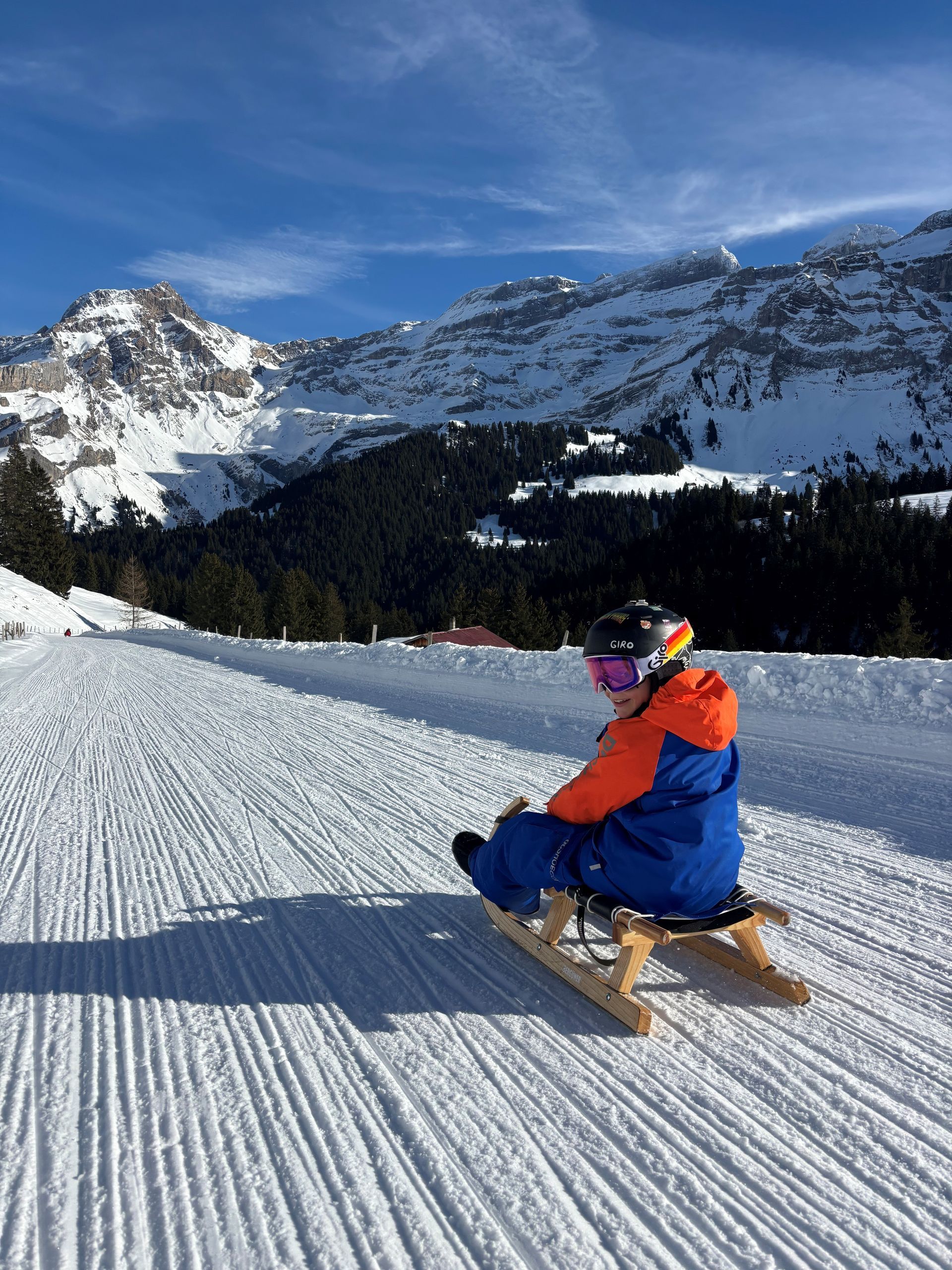 Skieur en veste orange et bleue assis sur une pente enneigée avec des sommets montagneux en arrière-plan