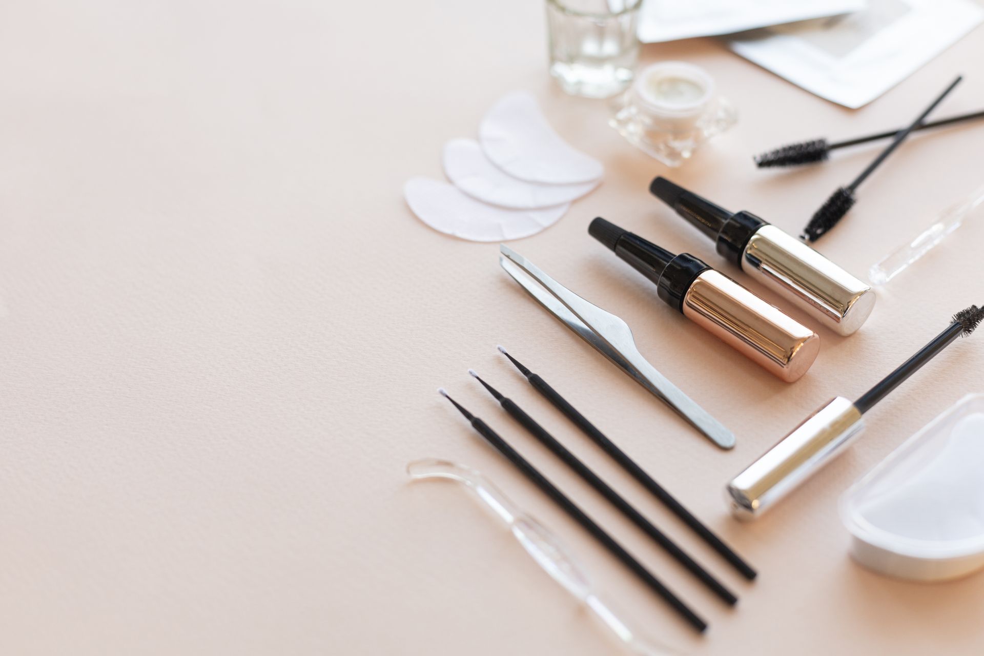 Beauty-Tools und -Produkte: Mascara, Bürsten, Pinzetten, Schablonen, Pads und Cremes auf einer hellrosa Oberfläche.