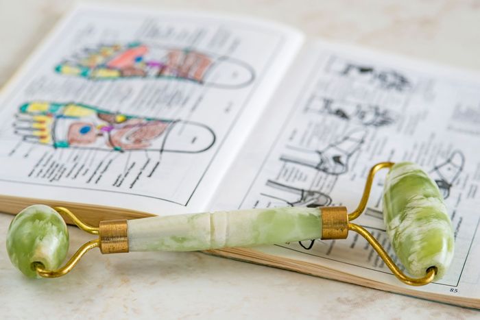 Jade-Gesichtsroller auf einem Buch mit Fußreflexzonenmassage-Diagrammen; goldene Beschläge, hellgrüner Stein.