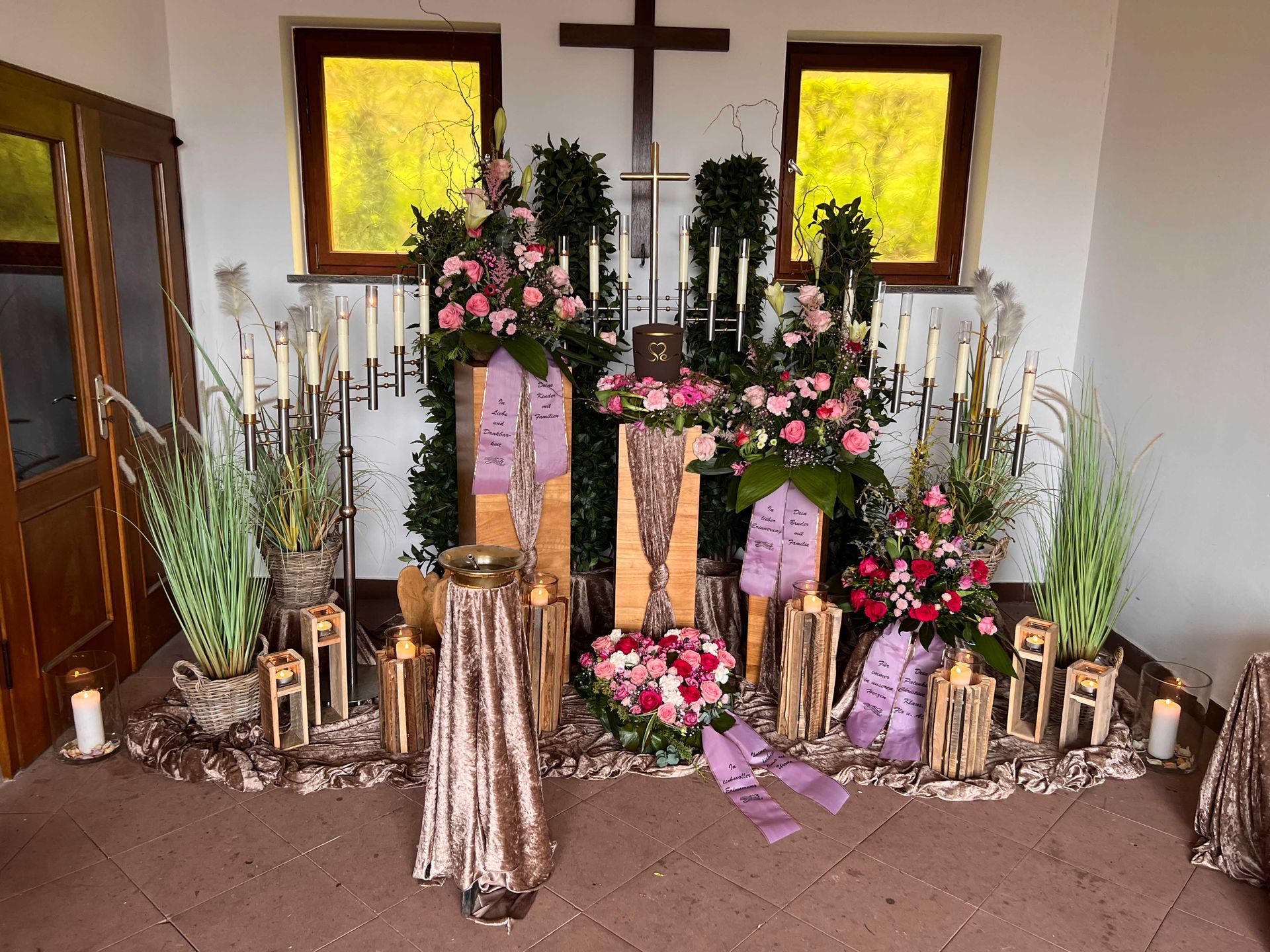 Grabgestaltung mit Blumen, Kerzen und Grablichtern vor Kreuz in einer Kapelle