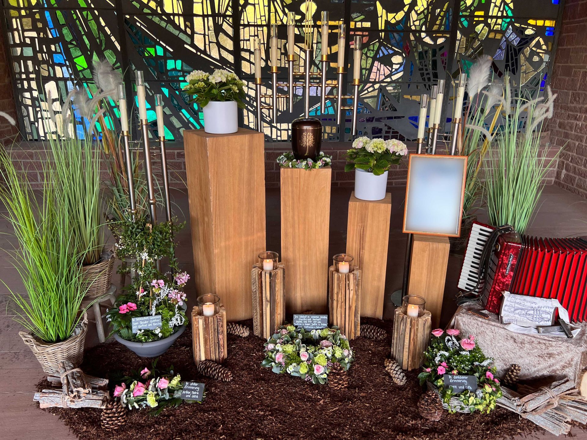 Grabgestaltung mit Holzurnen, Blumenschmuck, Grablichtern und Kerzen vor Kirchenfenster