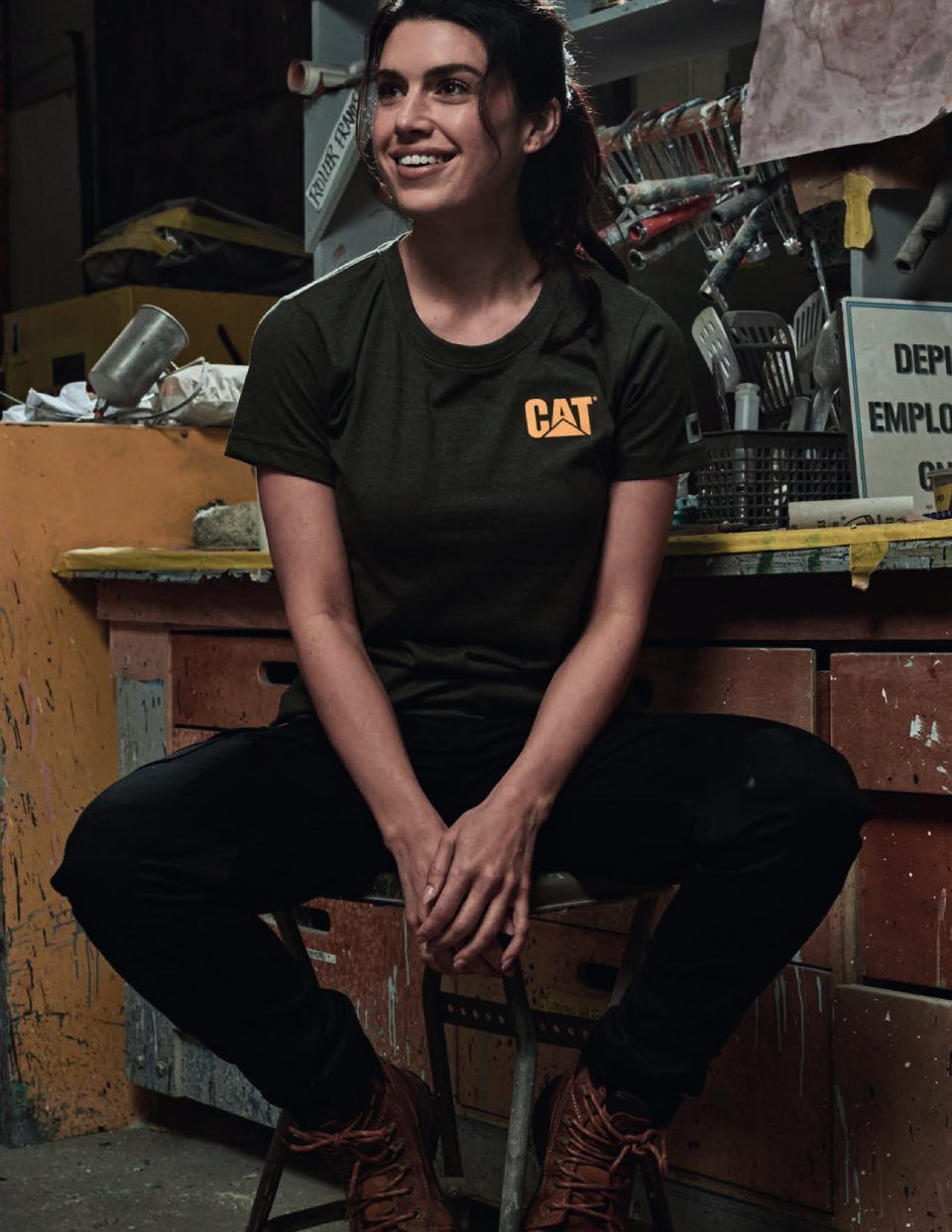 Eine Frau in einem Katzen-T-Shirt sitzt lächelnd mit übereinandergeschlagenen Beinen in einer Werkstatt.