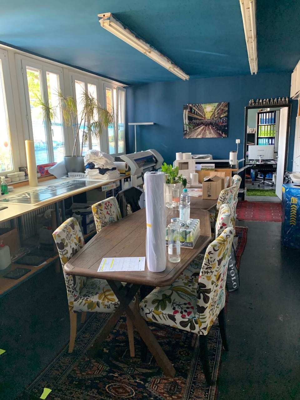 Innenansicht eines Zimmers mit einem langen Holztisch, umgeben von gemusterten Stühlen, mit blauer Decke und Wänden.