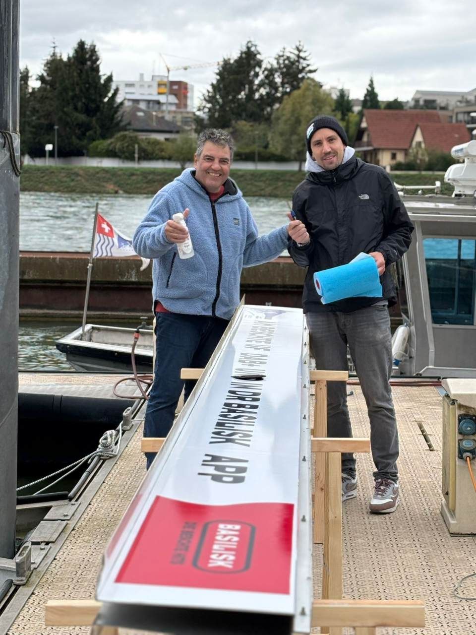 Zwei Männer halten an einem Dock ein langes Schild mit der Aufschrift „MISSION APP“. Einer zeigt mit dem Daumen nach oben, der andere deutet darauf.