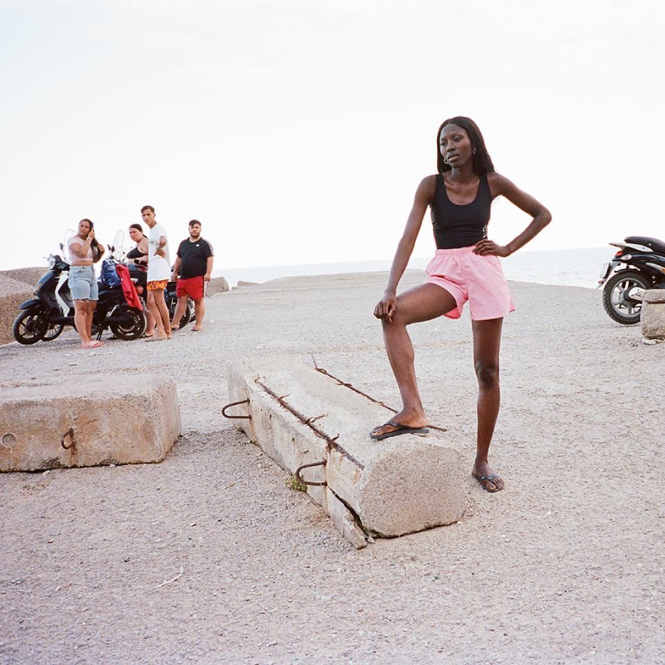 Eine Frau in pinkfarbenen Shorts und schwarzem Top steht auf einem Betonblock, andere stehen neben Rollern.