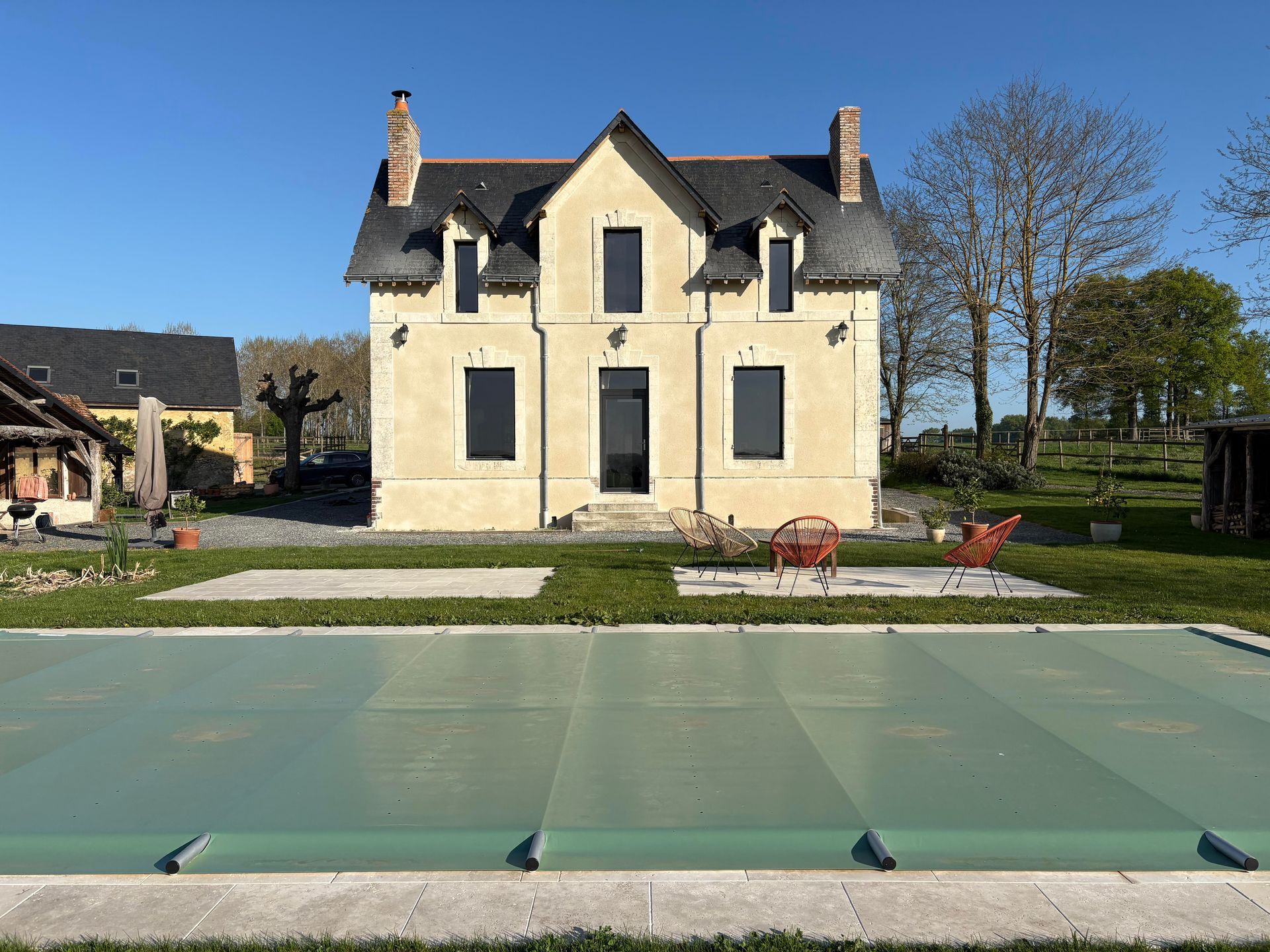 Maison de deux étages couleur crème avec une bâche de piscine verte à l'avant, sous un ciel bleu azur.
