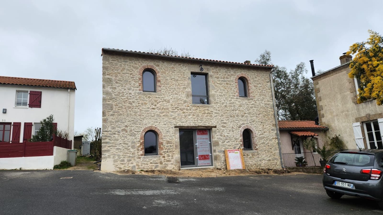 Bâtiment en pierre avec fenêtres cintrées et porte rouge, à côté de voitures garées dans une petite cour