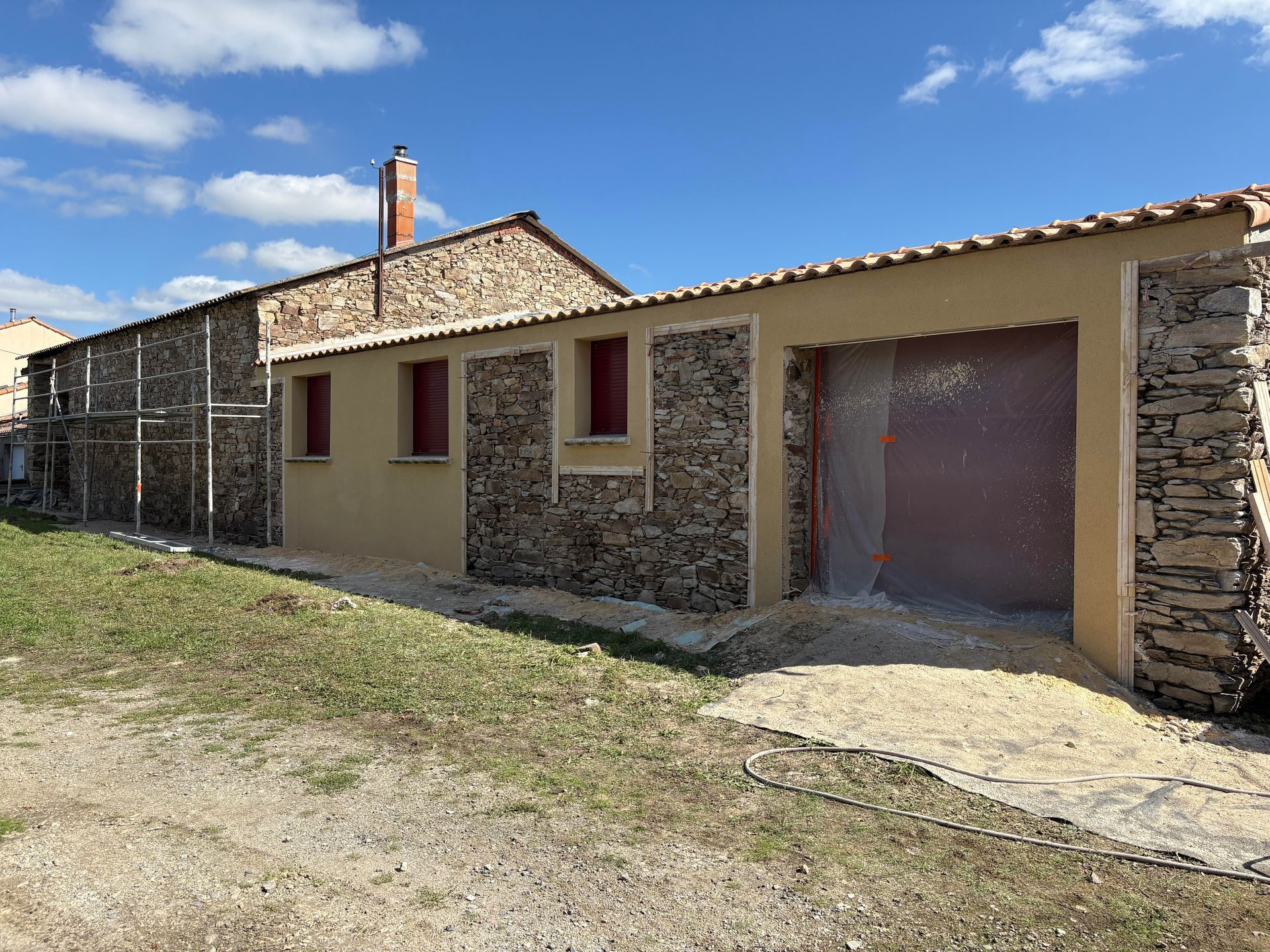 Ferme en pierre avec volets rouges et grande porte de garage sous un ciel bleu.