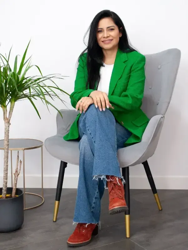 Una persona con cabello largo y oscuro está sentada en una silla gris, vistiendo un blazer verde brillante y una blusa blanca