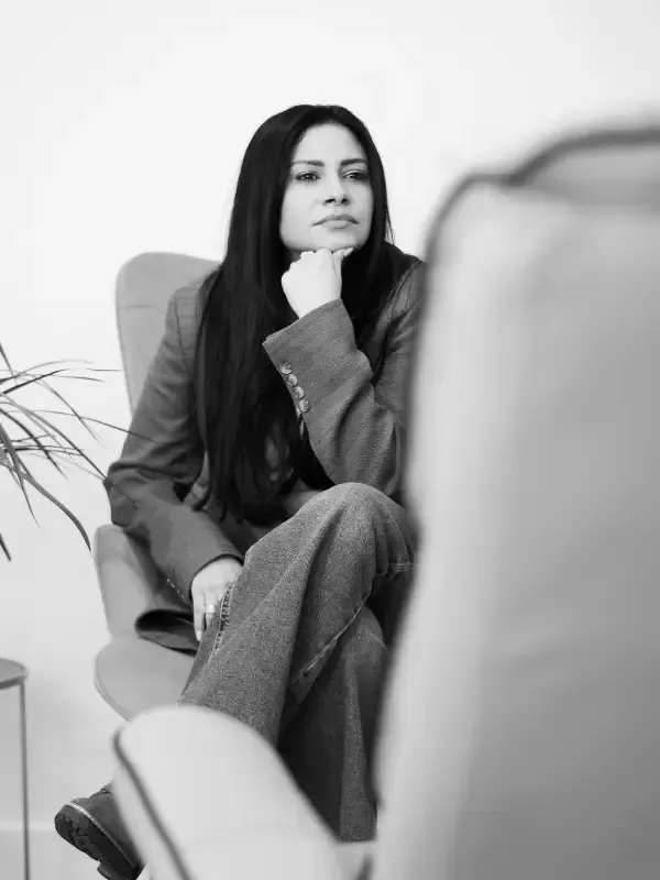 Una persona con cabello largo y oscuro está sentada en un sillón, apoyando la barbilla en la mano y mirando pensativamente