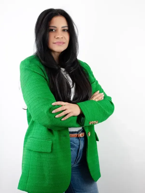 Una persona con cabello largo y oscuro, vestida con una chaqueta de tweed verde y jeans, permanece de pie