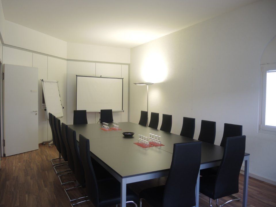 Konferenzraum mit langem Tisch, schwarzen Stühlen, Projektionsleinwand und Wandleuchte