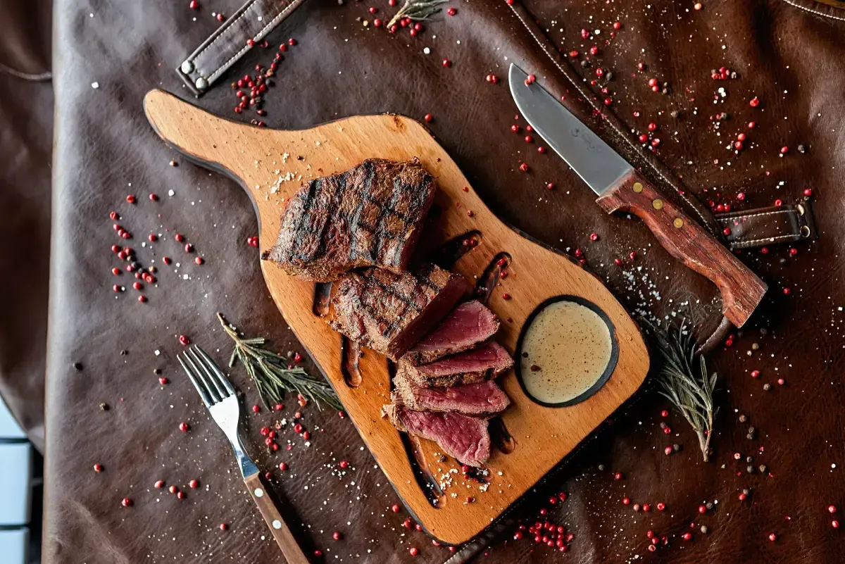 Un filete a la parrilla, cortado en rodajas, servido con salsa para mojar en una tabla de madera con tenedor, cuchillo