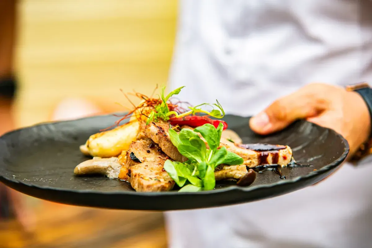 Una persona con uniforme blanco sostiene un plato negro con un plato gourmet adornado con verduras frescas.
