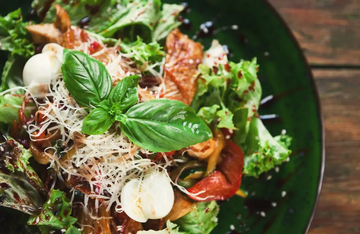Una ensalada gourmet en un plato verde oscuro, cubierta con hojas de albahaca fresca, queso rallado y perlas de mozzarella.