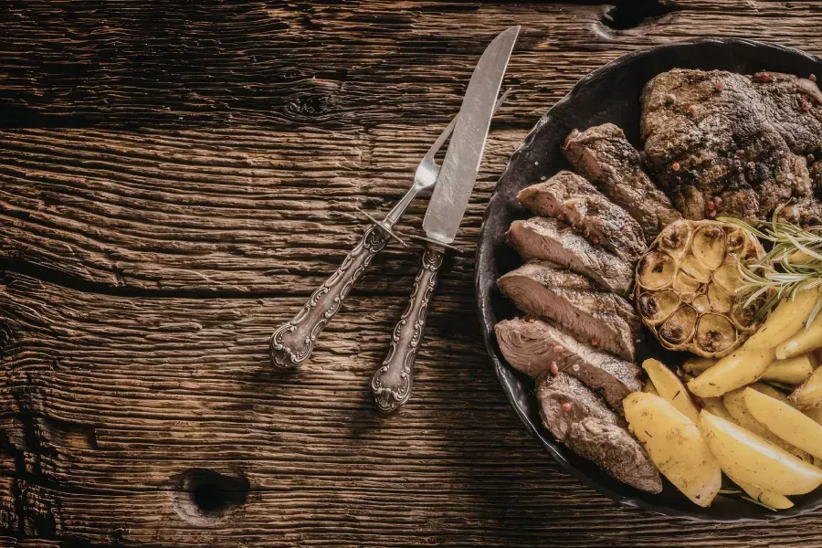 Una sartén de hierro fundido con filete en rodajas, ajo asado y patatas gajo, junto a un cuchillo 