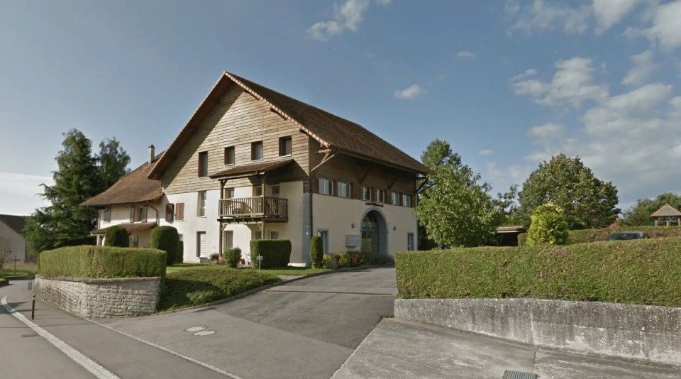 Une grande maison blanche avec un toit marron est entourée d'arbres et de buissons.