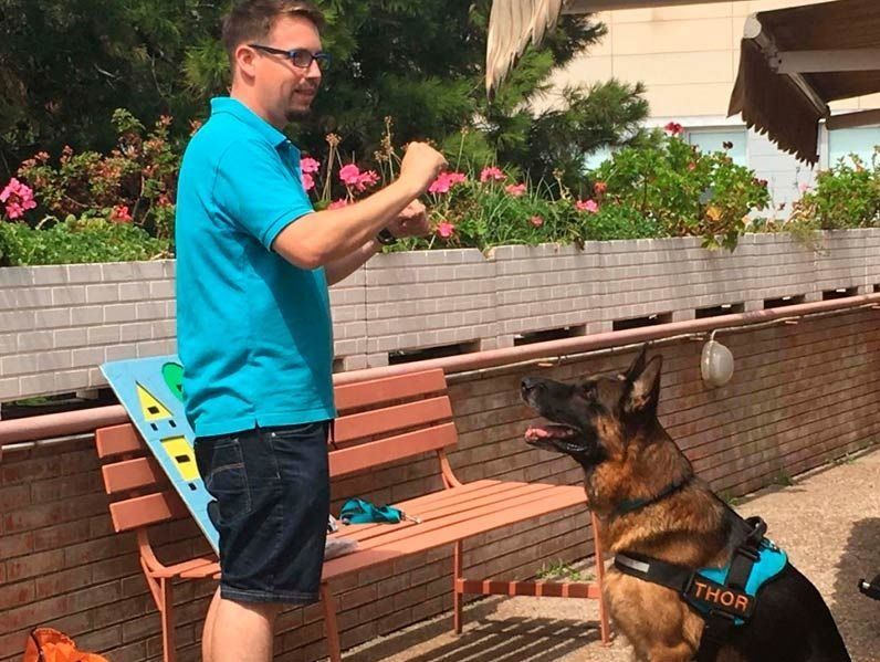 Un home està dret al costat d'un pastor alemany en un banc.