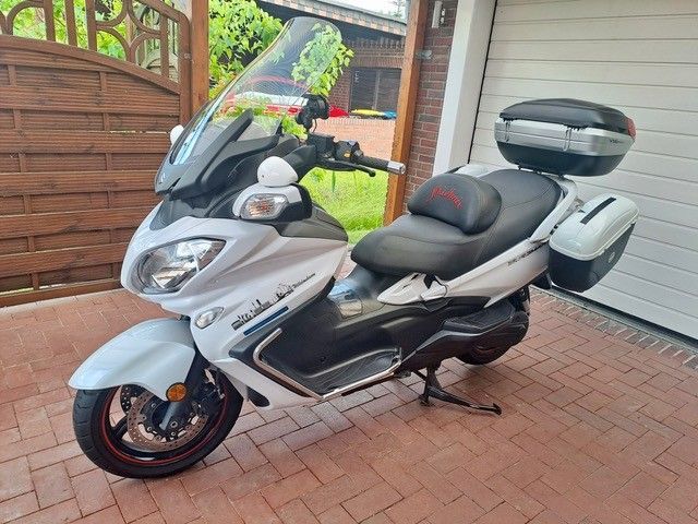 Weißer Motorroller mit schwarzem Sitz, Gepäckraum hinten und Gepäckraum an der Seite, geparkt auf einer gepflasterten Auffahrt neben einer Garage.