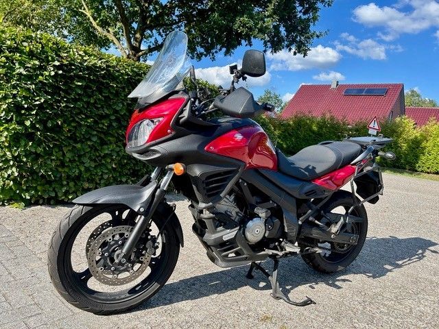 Ein rot-schwarzes Motorrad parkt auf einer sonnigen Straße; im Hintergrund sind Bäume und eine Windschutzscheibe zu sehen.