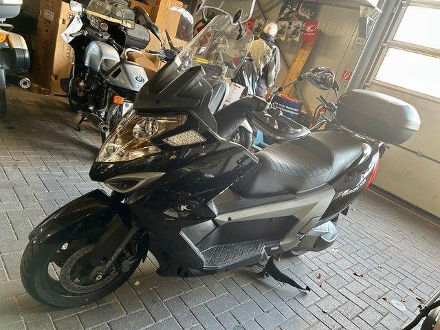 Ein schwarzer Roller steht in einem Innenraum auf einem Ziegelboden; im Hintergrund sind weitere Motorräder zu sehen.